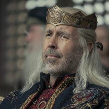 A man wearing a crown sits in a chair surrounded by others.